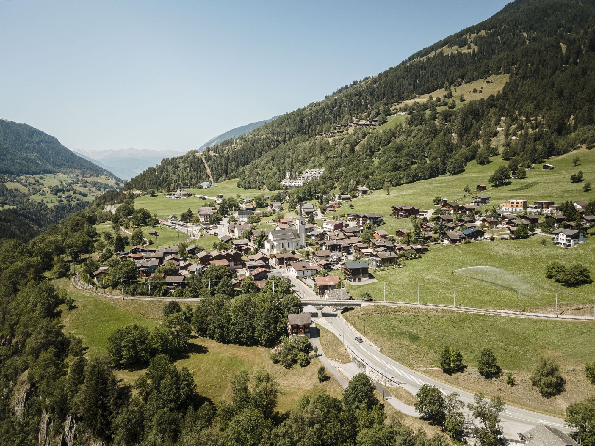 Blick auf ein idyllisches Bergdorf mit Kirche im Zentrum, umgeben von traditionellen Häusern, Bäumen und grünen Berghängen unter strahlend blauem Himmel.